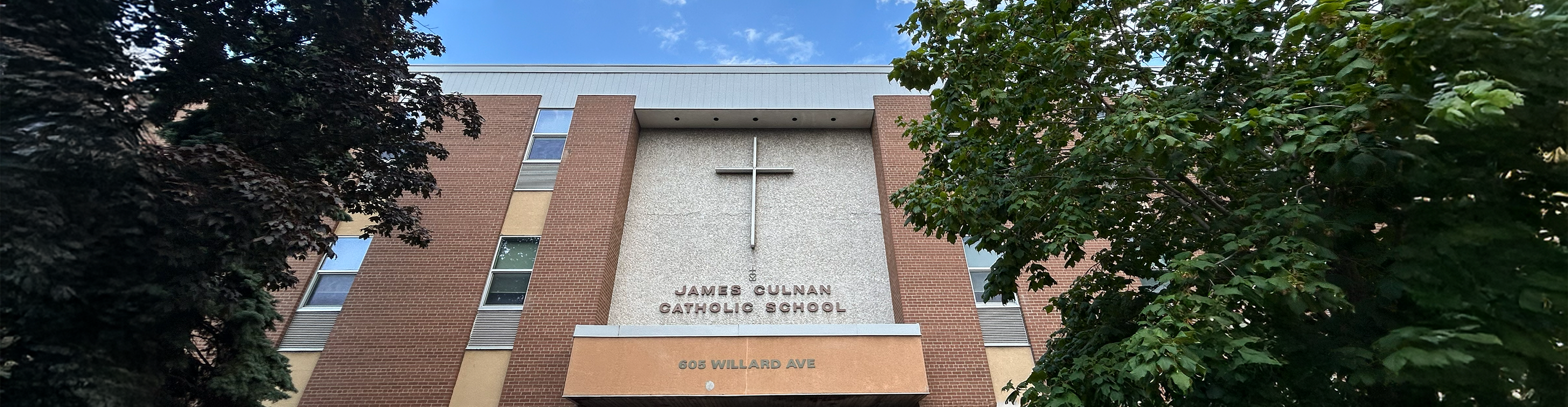 Photo of the front of the James Culnan Catholic School building, showing the beige stone and red brick building nestled between trees, with the school name and a large cross on the front