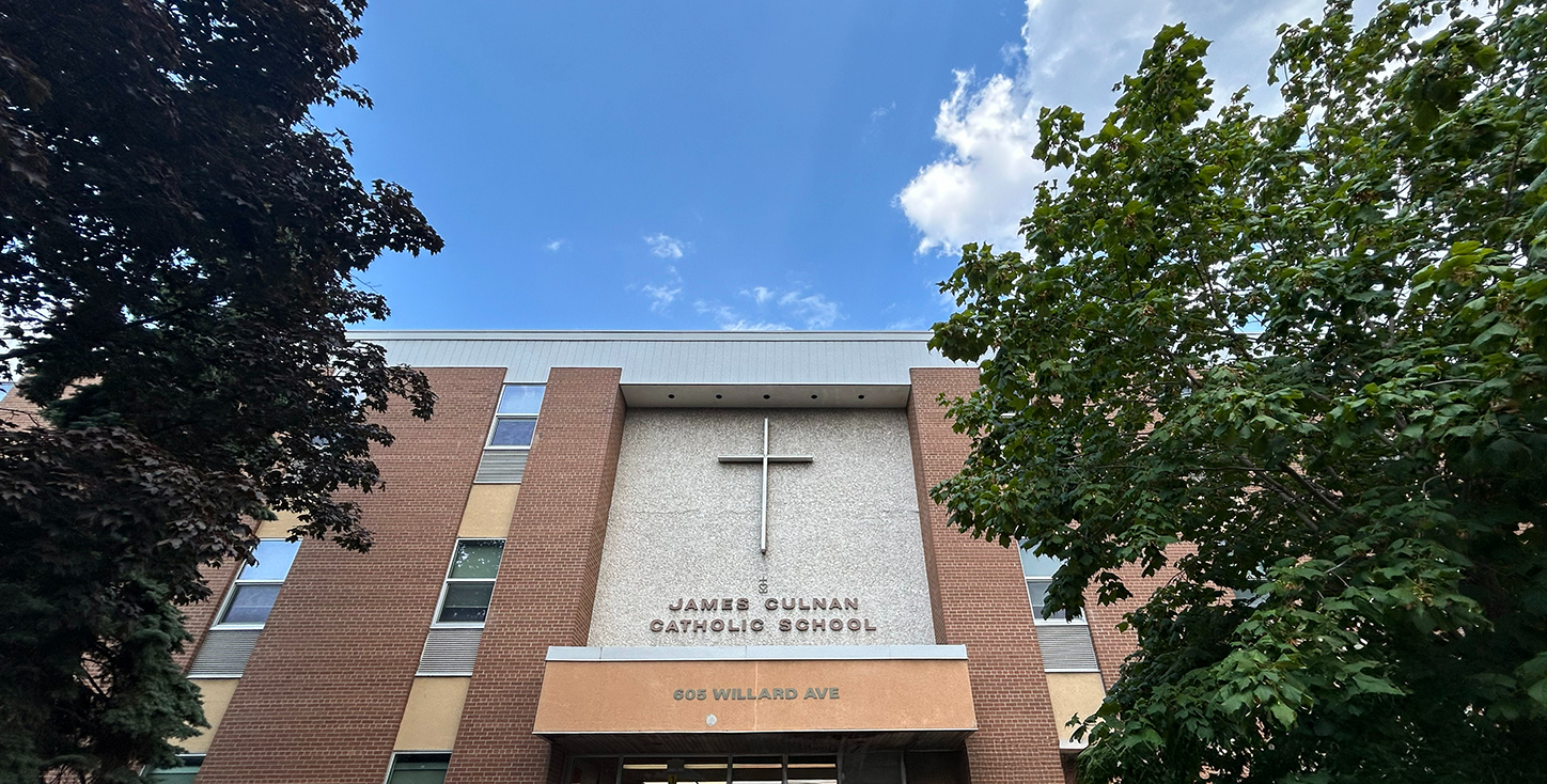Photo of the front of the James Culnan Catholic School building, showing the beige stone and red brick building nestled between trees, with the school name and a large cross on the front