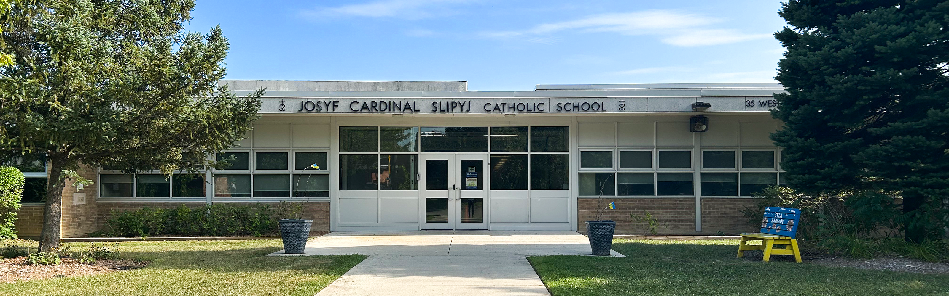 Photo of the Josyf Cardinal Slipyj Catholic School building, showing the white stone building from the front, bordered by trees, with the school name in black above the front door