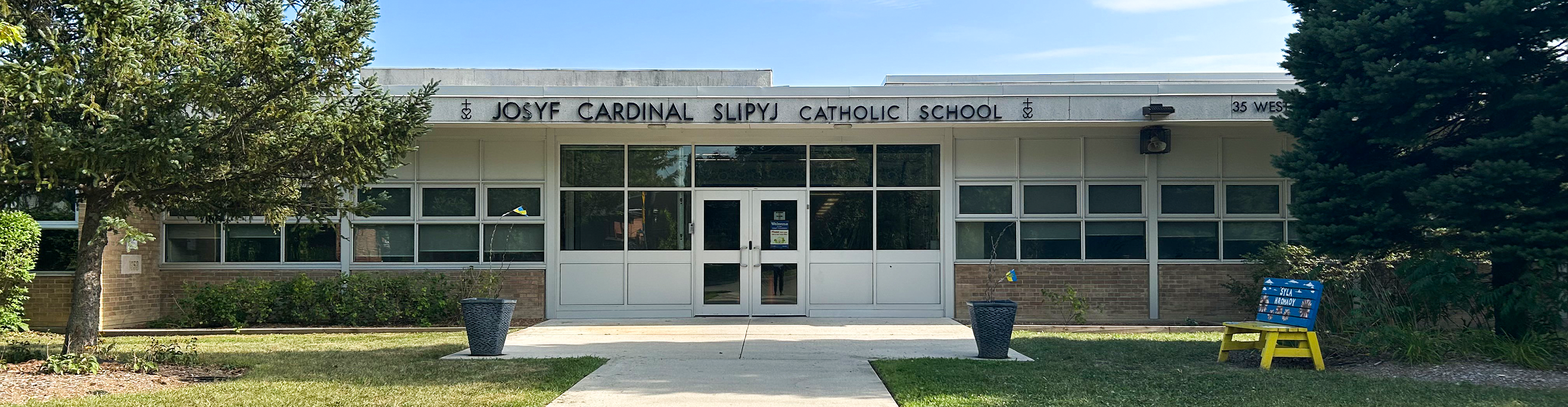 Photo of the Josyf Cardinal Slipyj Catholic School building, showing the white stone building from the front, bordered by trees, with the school name in black above the front door