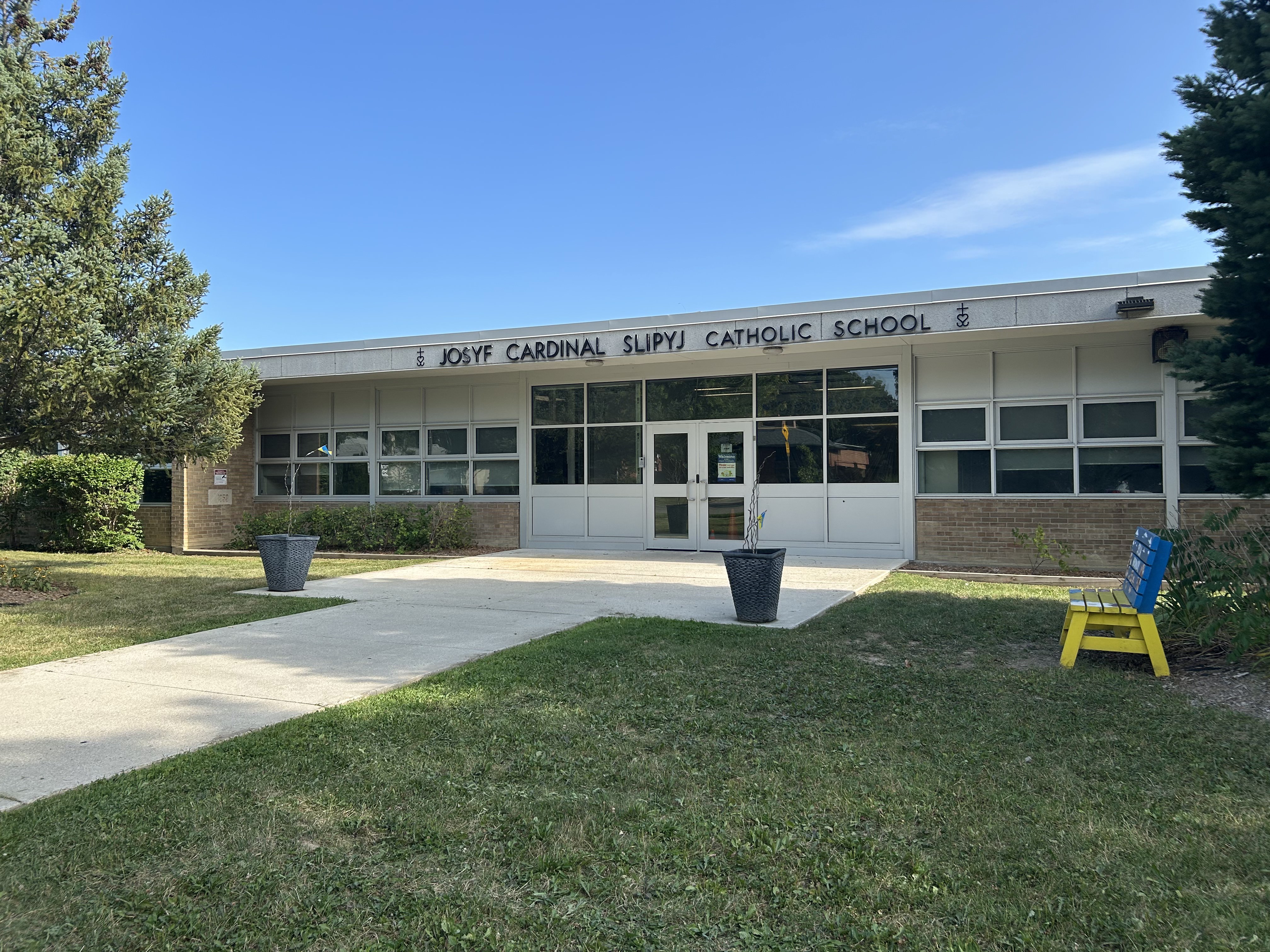 Photo of the Josyf Cardinal Slipyj Catholic School building, showing the white stone building from the front, bordered by trees, with the school name in black above the front door