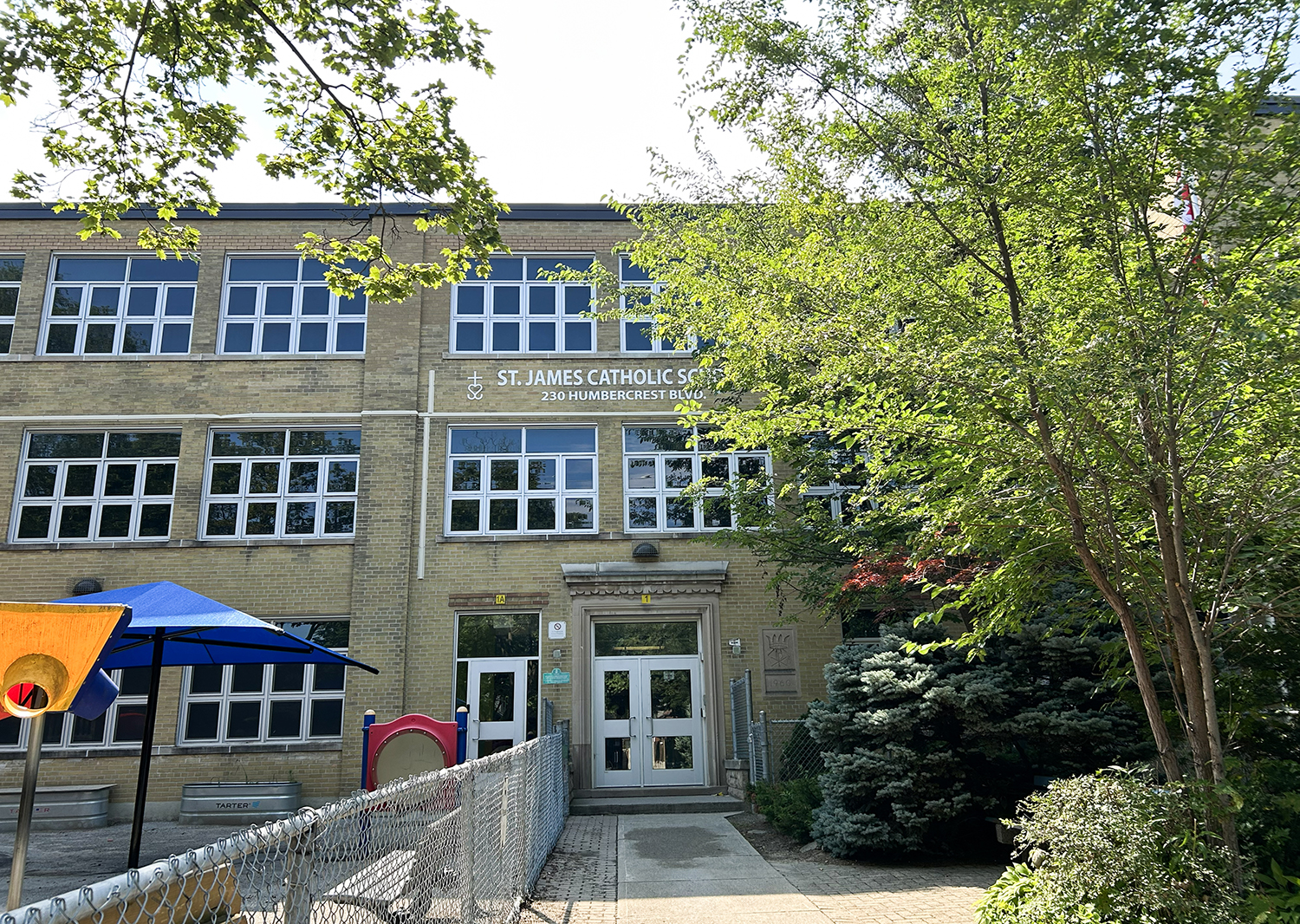 The front of the St. James Catholic School building, showing a beige brick building bordered by tall trees and looking out onto a residential road