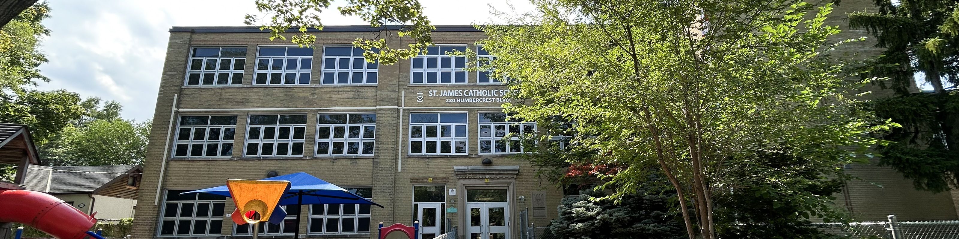 The front of the St. James Catholic School building, showing a beige brick building bordered by tall trees and looking out onto a residential road