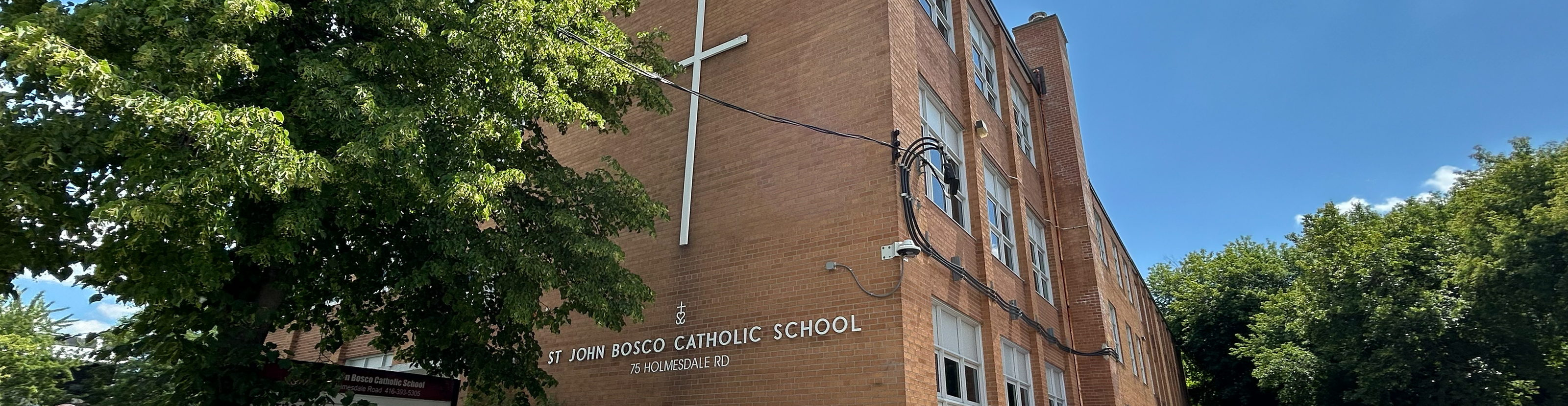 The front of the St. John Bosco Catholic School building, showing a red brick building surrounded by tall trees and greenery, with the school name in white on the front, along with a large cross above the name
