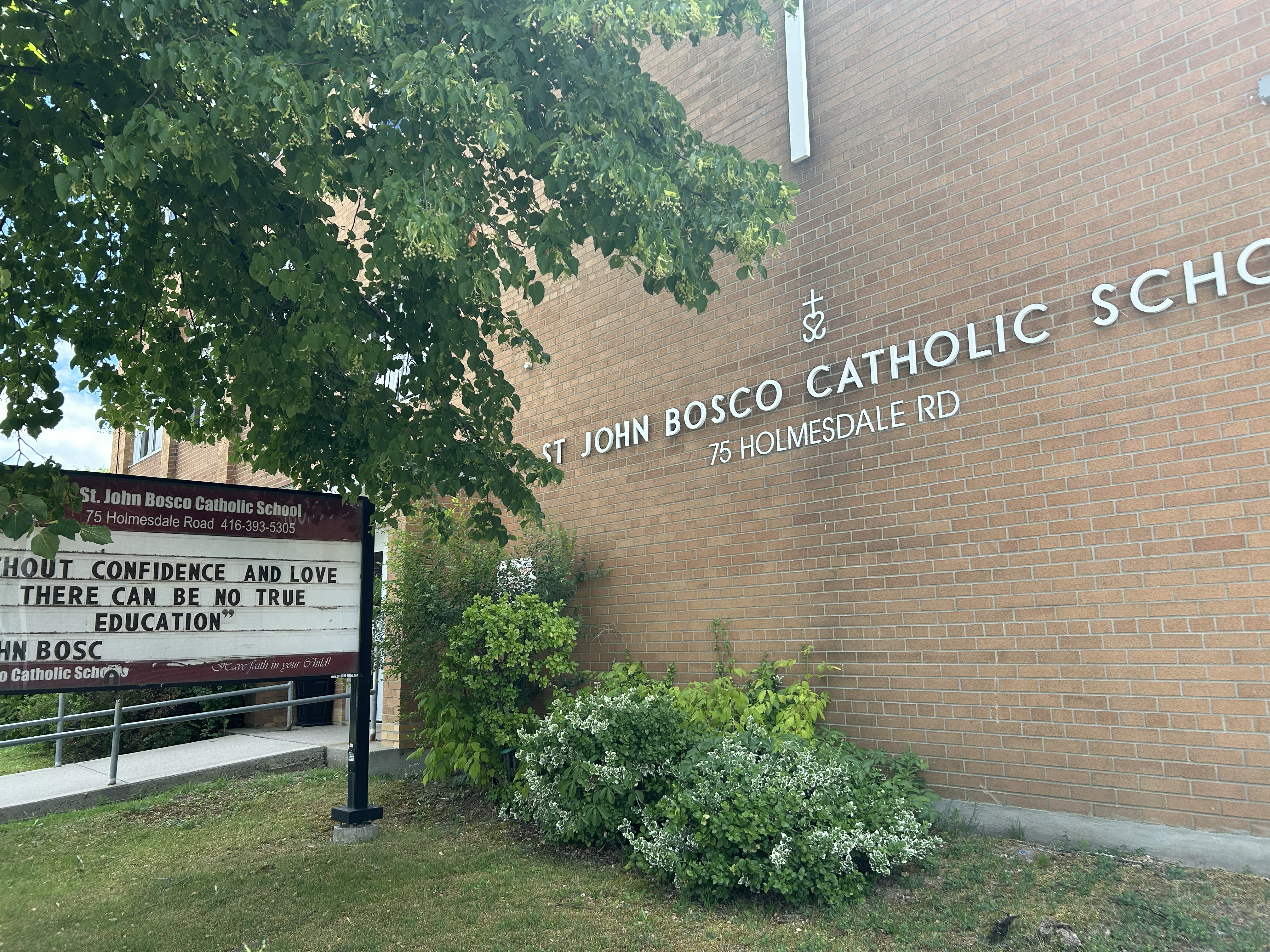The front of the St. John Bosco Catholic School building, showing a red brick building surrounded by tall trees and greenery, with the school name in white on the front, along with a large cross above the name