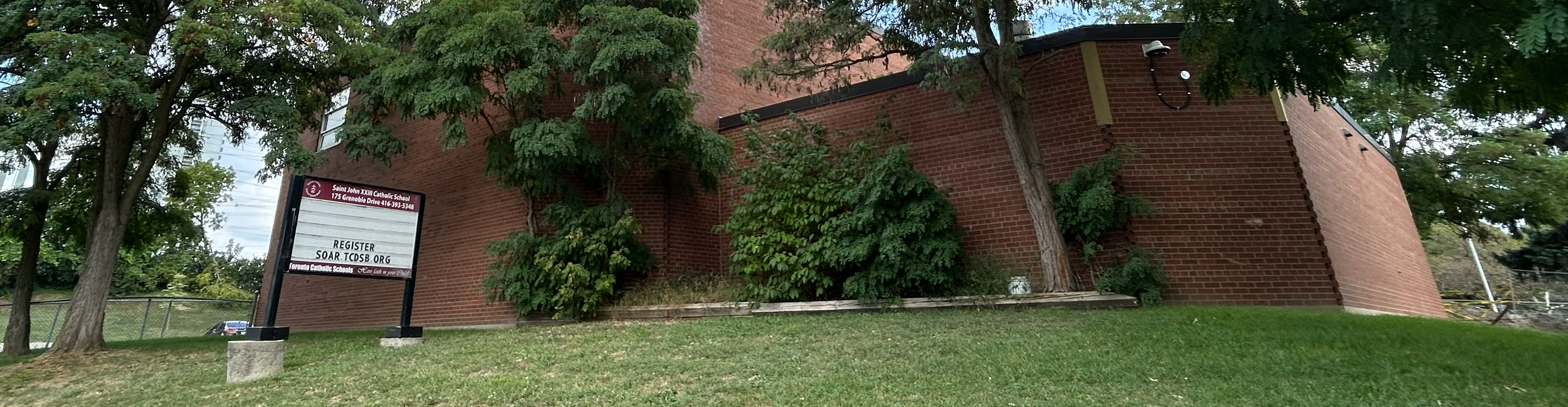 A photo of the St. John XXIII Catholic School building as seen from the road, showing a dark red brick building nestled between tall trees and greenery