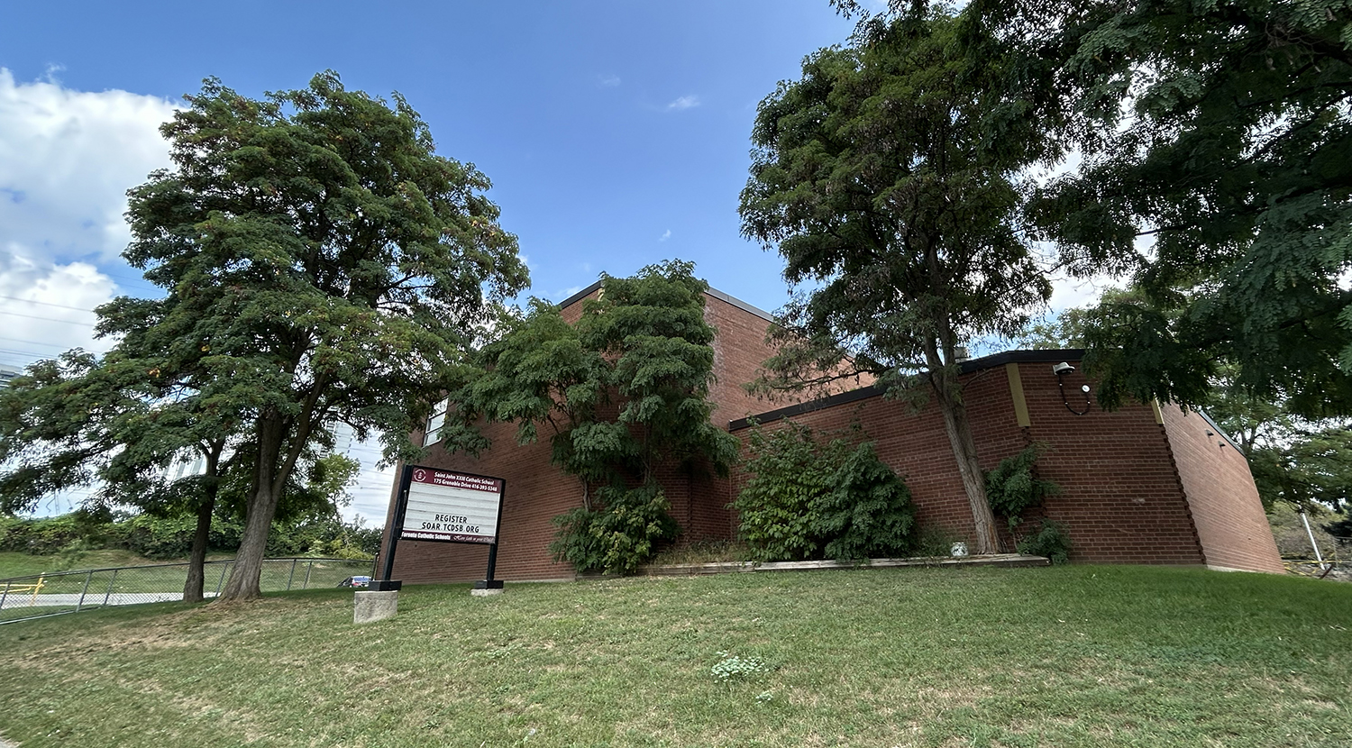 A photo of the St. John XXIII Catholic School building as seen from the road, showing a dark red brick building nestled between tall trees and greenery