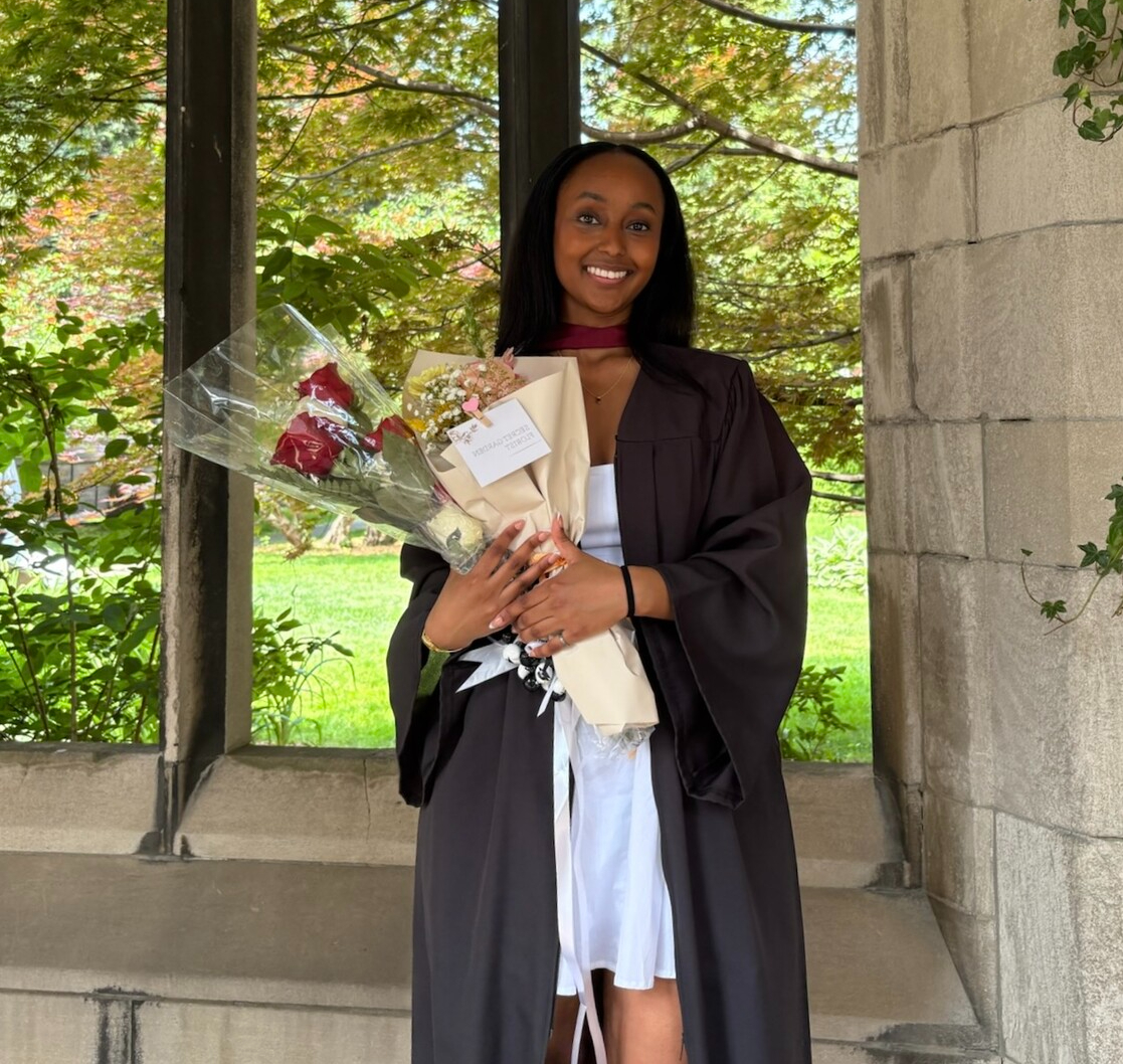Photo of Dina in her graduation robes, holding congratulatory bouquets of flowers