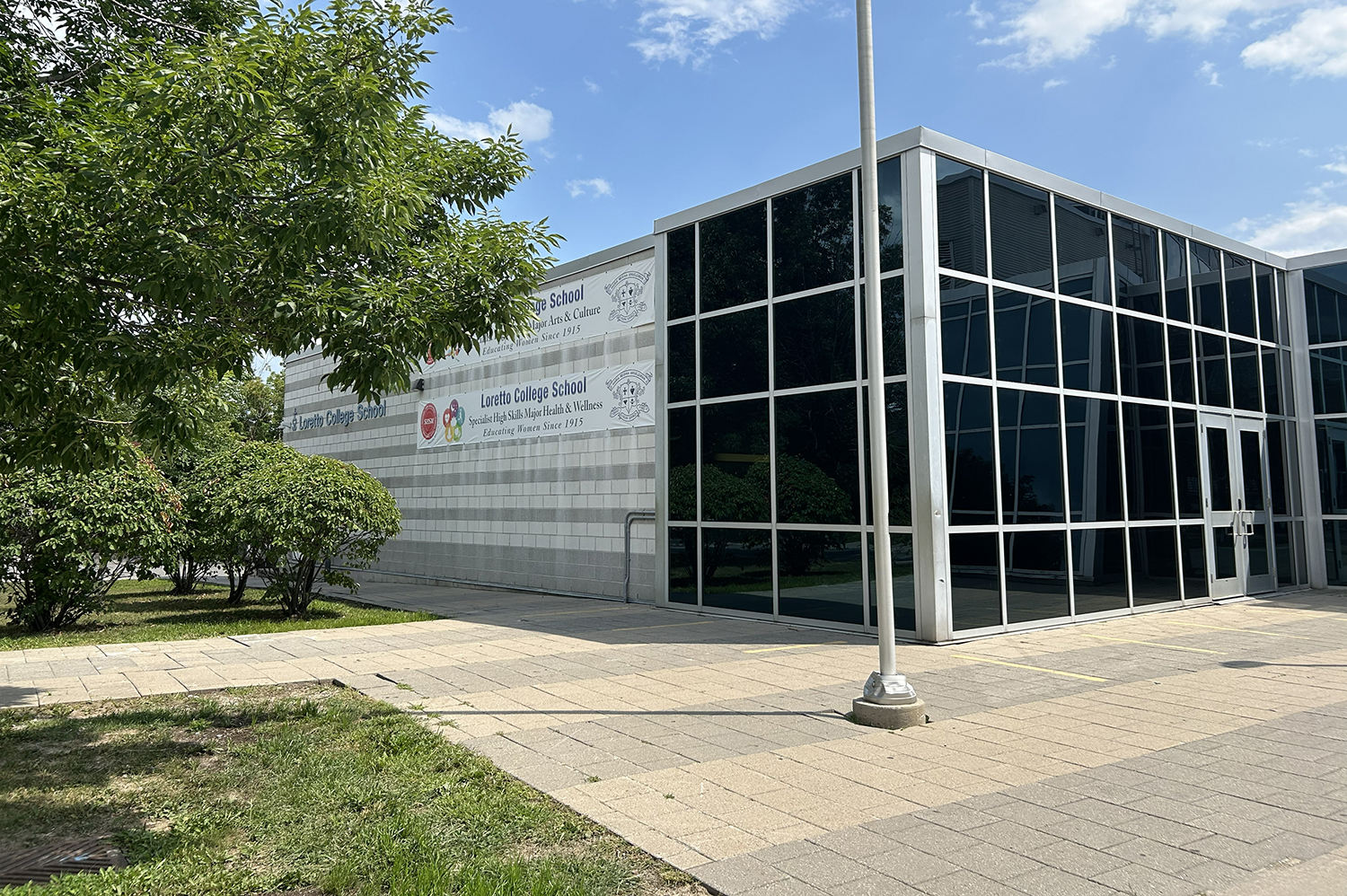 The front entrance of the Loretto College School, showing a white and glass walled modern-looking building situatd among tall and verdant trees