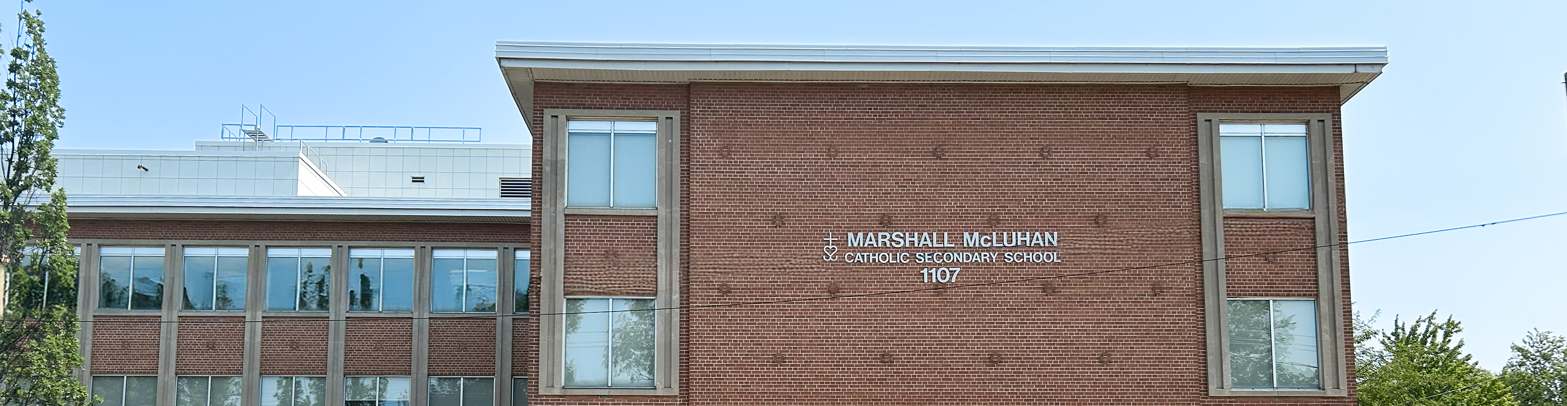 Photo of the Marshall McLuhan Catholic Secondary School building, showing a light red brick building along the side of the road with trees around it and the school name on the front