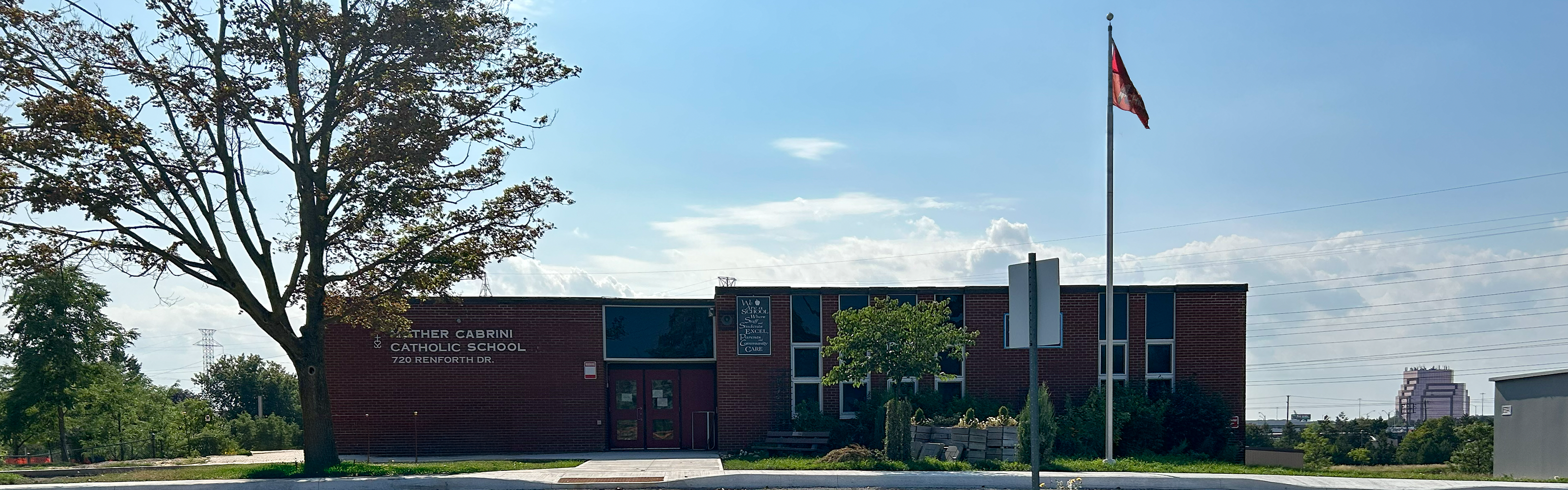 The front of the Mother Cabrini Catholic School building, showing the red brick building against a sunny blue sky with expansive green ground beyond