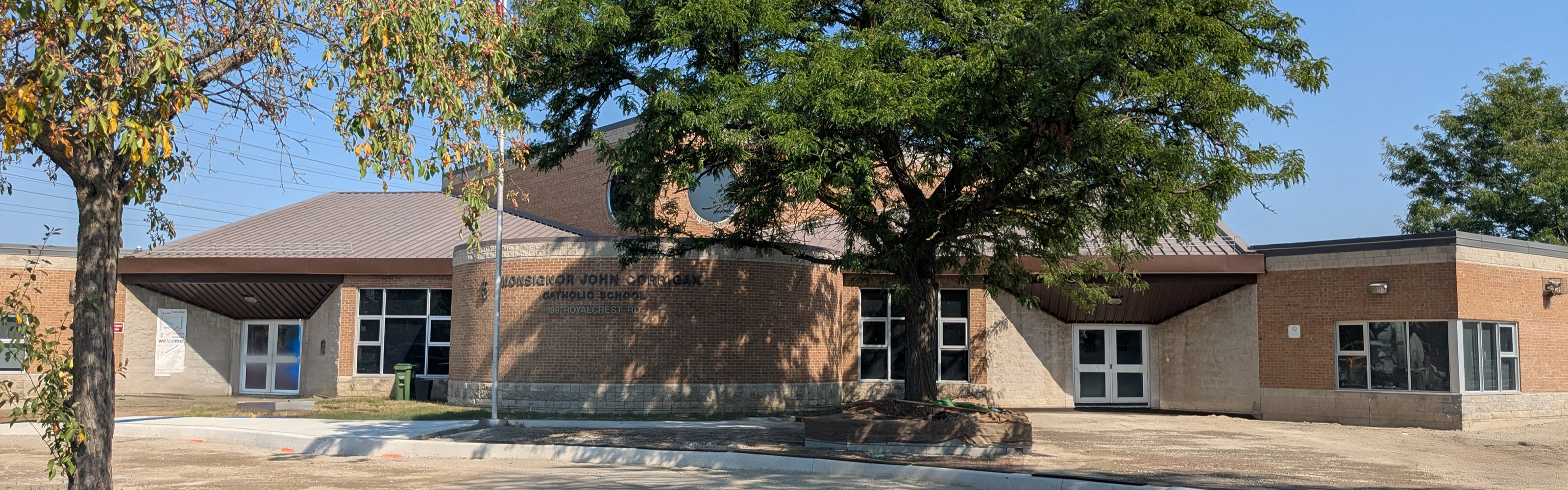 A photo of the Monsignor John Corrigan Catholic School building, showing a uniquely-shaped tan brick building displaying the school name in dark brown, nestled between trees.
