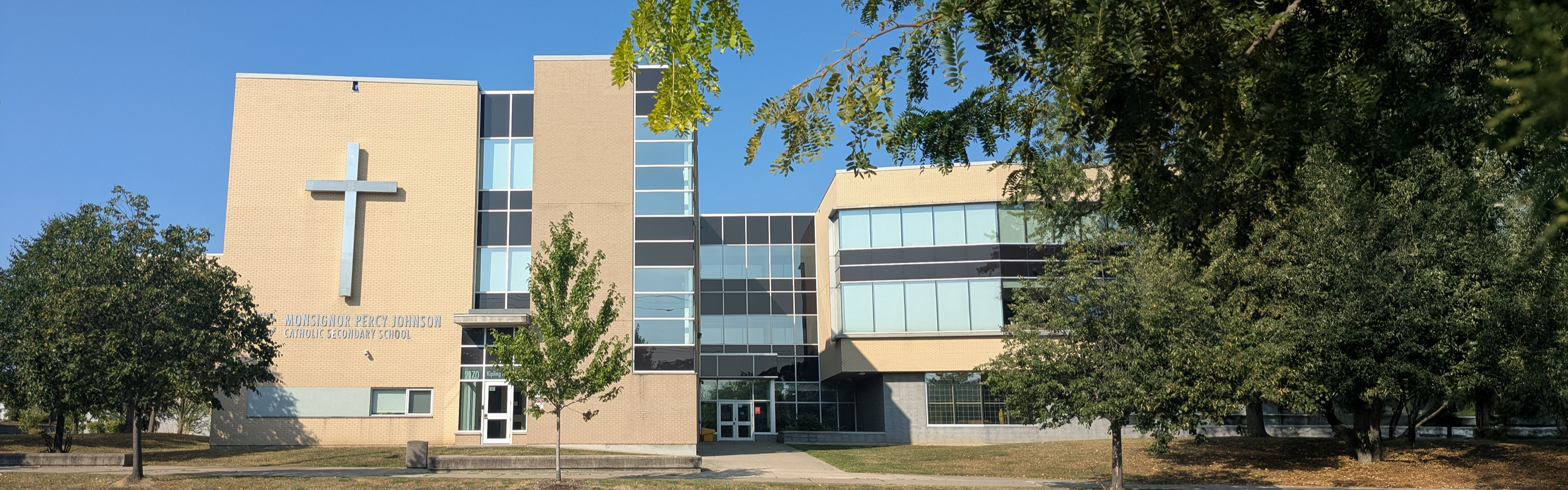 A photo of the Monsignor Percy Johnson Catholic Secondary School building, showing a modern-style pale brick building displaying the school name and a large cross in silver, with large alternating clear and tinted windows nestled between trees and looking onto the street. 