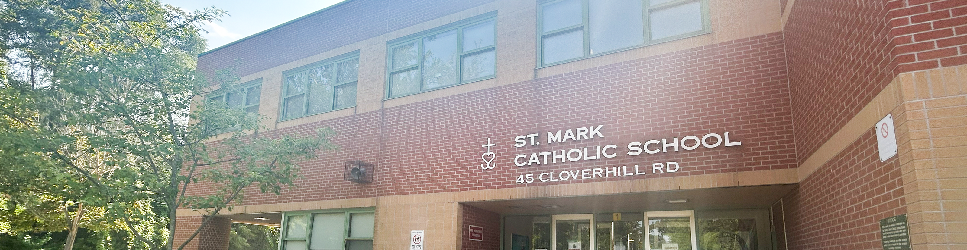 The front of the St. Mark Catholic School building, showing a dark red and orange brick building with the school playground in front and trees around, and the school name in white on the front along with the street address