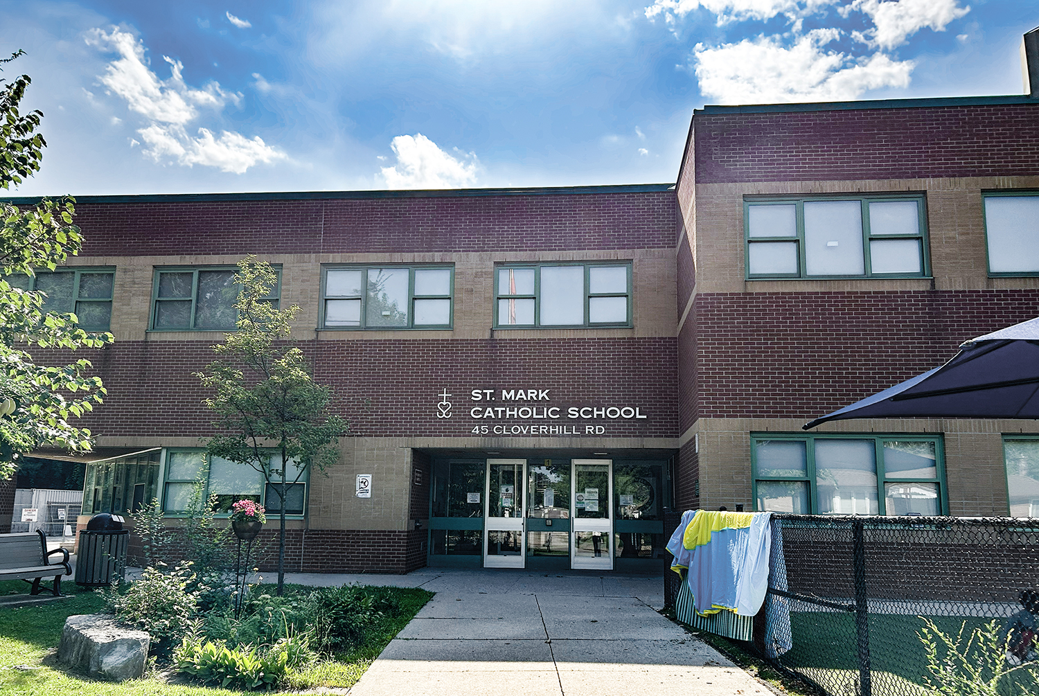 The front of the St. Mark Catholic School building, showing a dark red and orange brick building with the school playground in front and trees around, and the school name in white on the front along with the street address