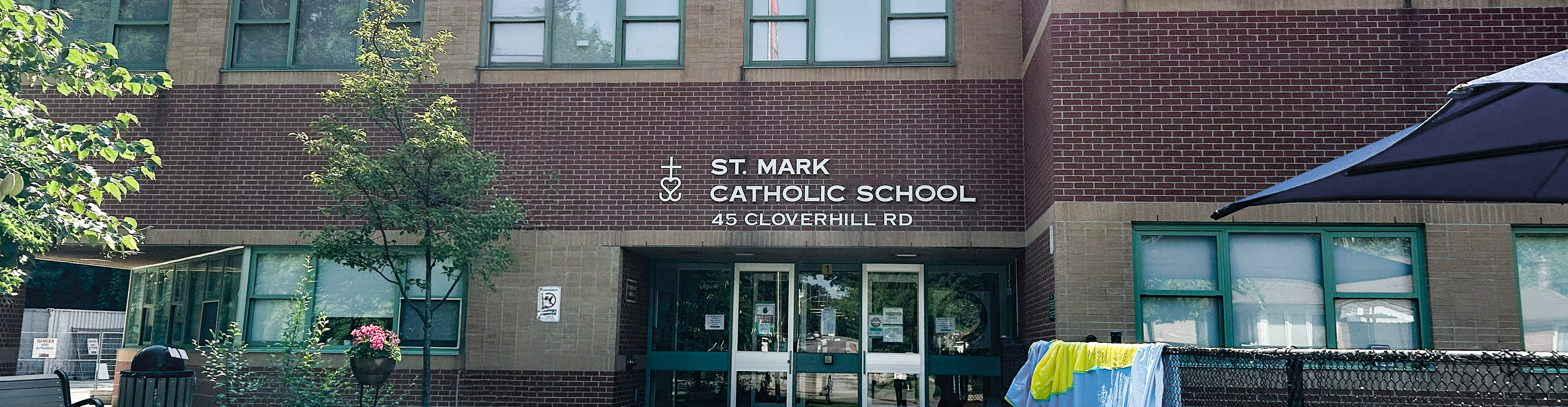 The front of the St. Mark Catholic School building, showing a dark red and orange brick building with the school playground in front and trees around, and the school name in white on the front along with the street address