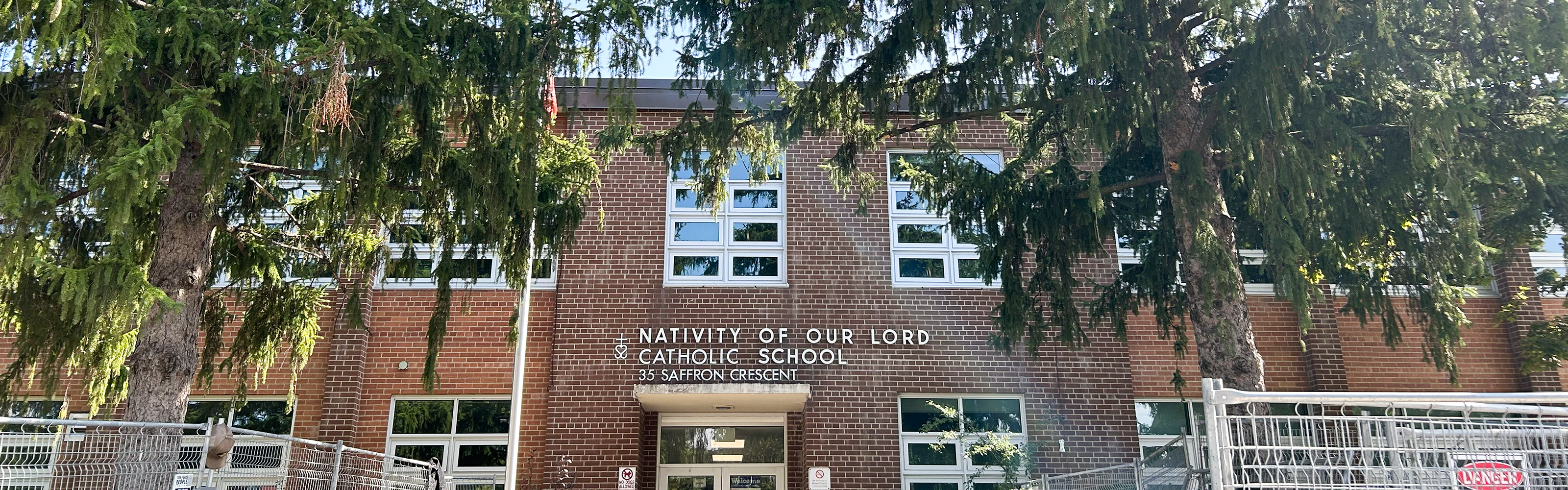 Photo of the Nativity of Our Lord Catholic School building, showing the red brick building nestled between tall trees, with the school name and address in white above the front door