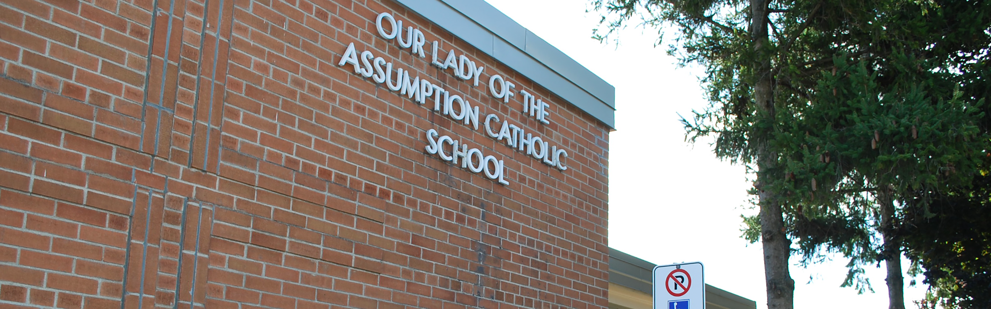 Photo of the Our Lady of The Assumption Catholic School building, showing a red brick building with the school name displayed in white on the wall, next to a large cross made from brick, beside some trees and an accessible parking sign.