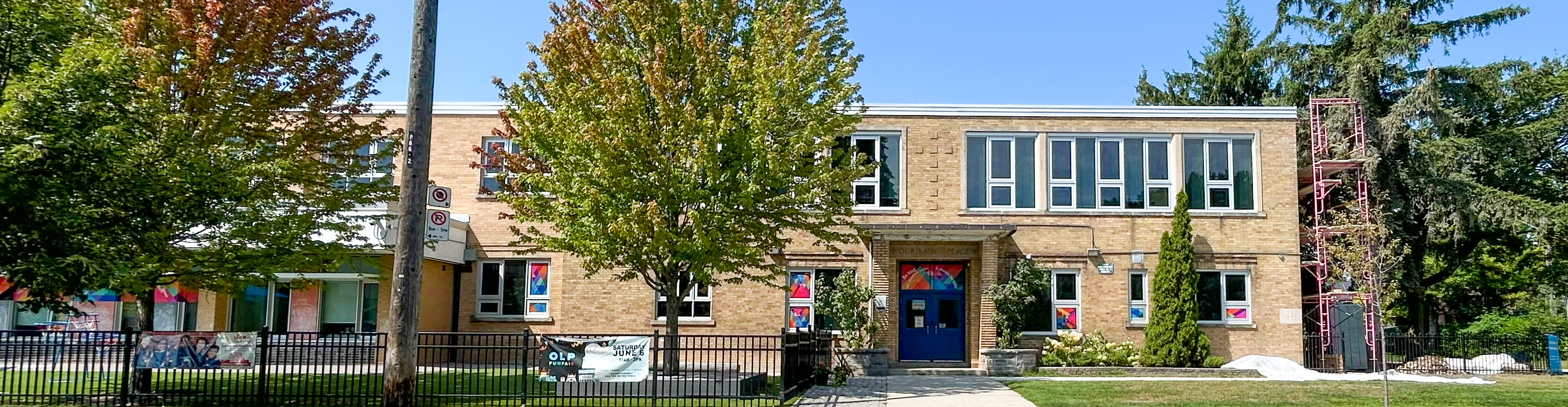 Photo of the front of the Our Lady of Peace building, showing a beige stone building bordered by tall green trees, looking out onto the road