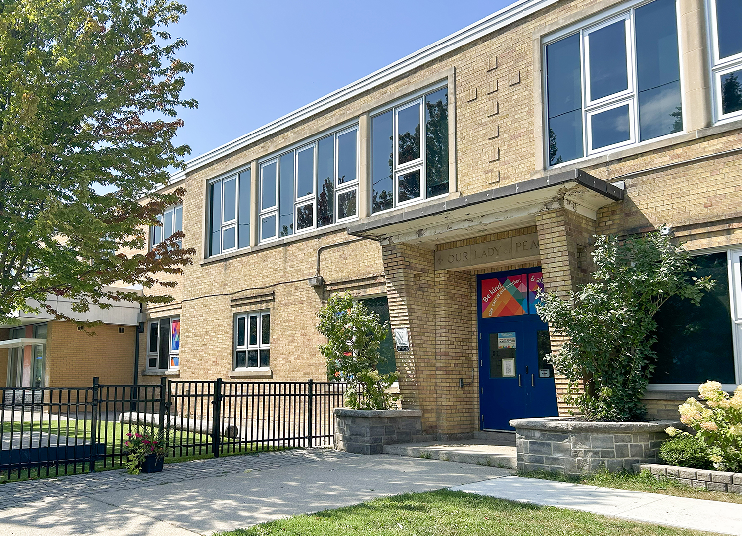Photo of the Our Lady of Peace Catholic School building, showing a beige brick building with blue double doors at the front looking out onto a residential street