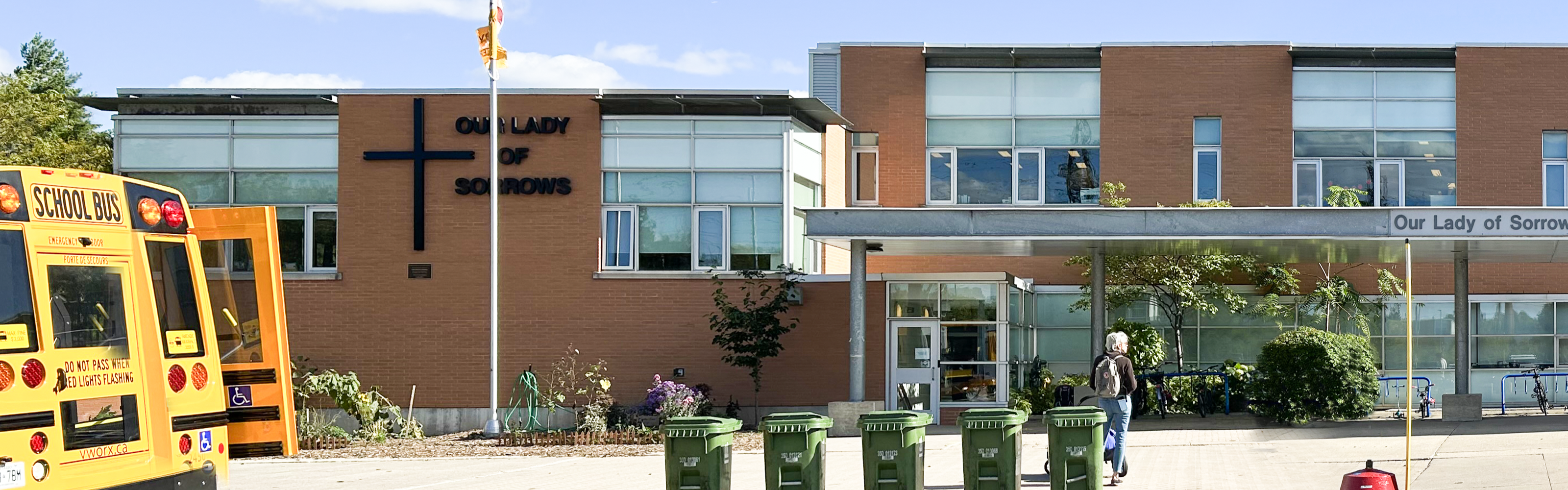 Photo of the Our Lady of Sorrows Catholic School building, showing a modern-style tan brick building with many windows, a prominent black cross and school name displayed on the front, a covered entrance, and a school bus parked in front.