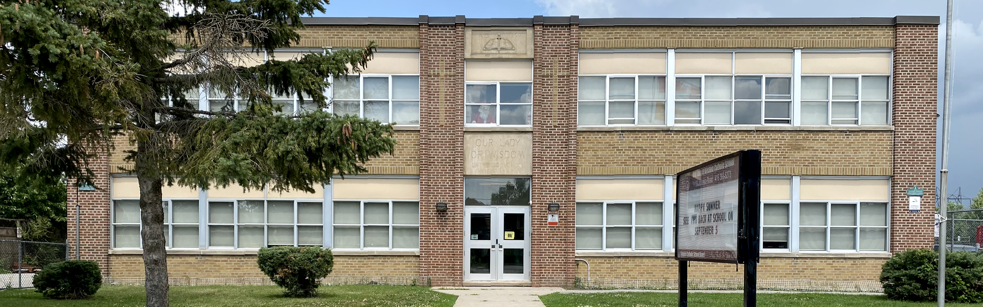 Photo of the Our Lady of Wisdom Catholic School building, showing classical-style tan brick building with red brick columns and white windows, the school name is inscribed above the entrance door, which display a cross shape within the columns framing the entrance. A large tree, bushes, grass, and fencing surround the building. 