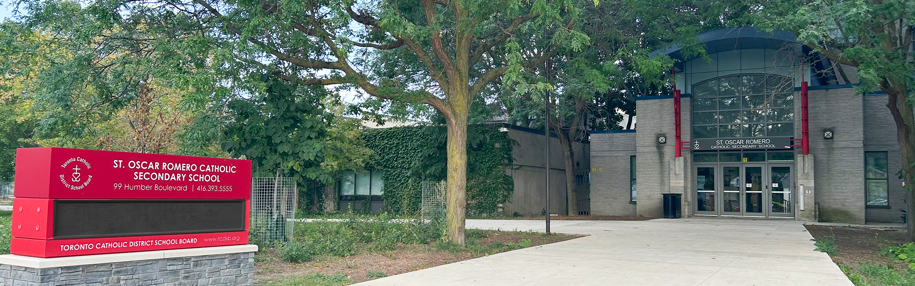 Photo of the front entrance of the St. Oscar Romero Catholic Secondary School building, showing the light grey stone building nestled between trees, with a red school sign posted outside near the front walkway