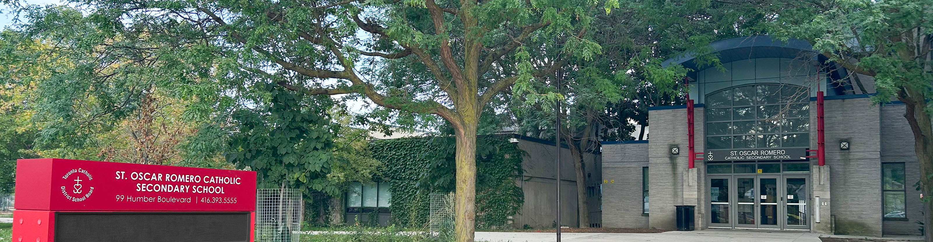 Photo of the front entrance of the St. Oscar Romero Catholic Secondary School building, showing the light grey stone building nestled between trees, with a red school sign posted outside near the front walkway