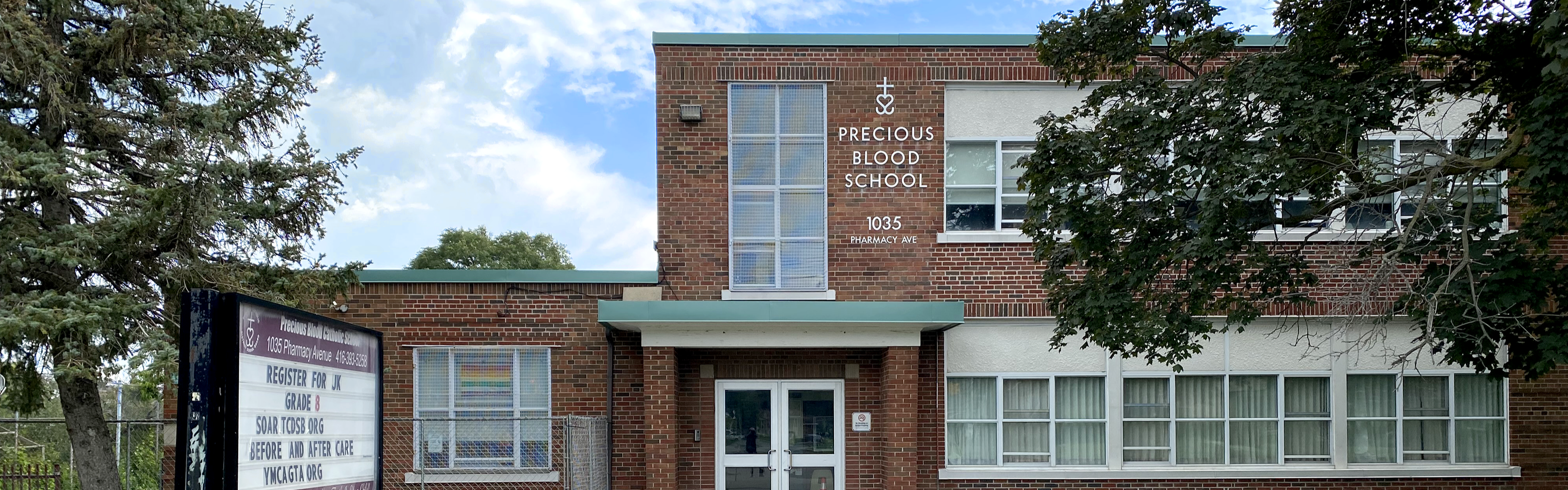 Photo of the Precious Blood Catholic School building, showing a red brick building with white windows and green paneling along the roof, the school name displayed in white, and the school sign to the left of the entrance, surrounded by trees. 