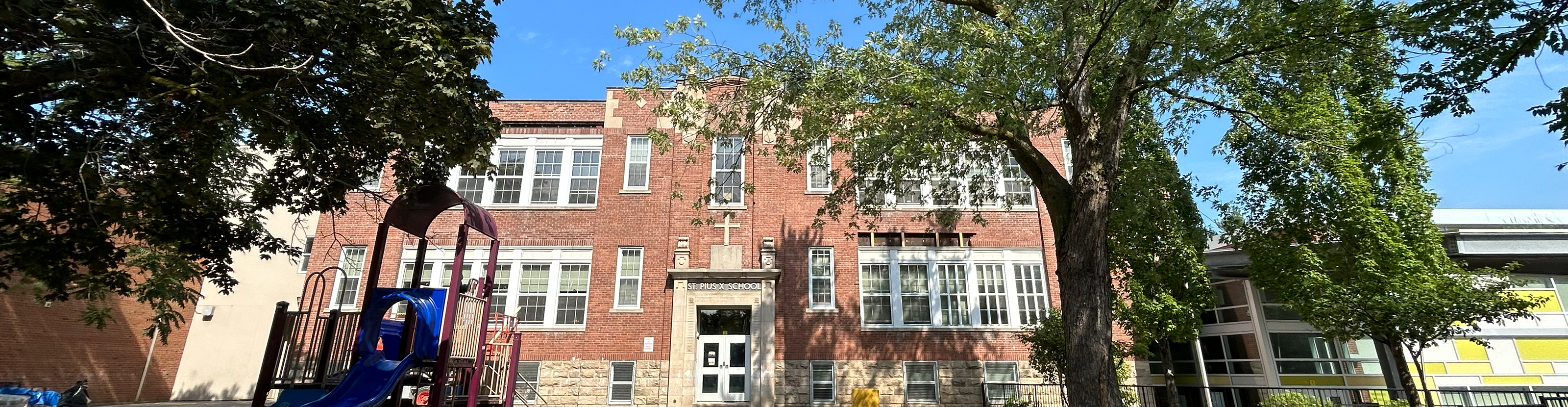 The front of the St. Pius X Catholic School building, showing a light brown building with the school play ground in front and tall trees bordering