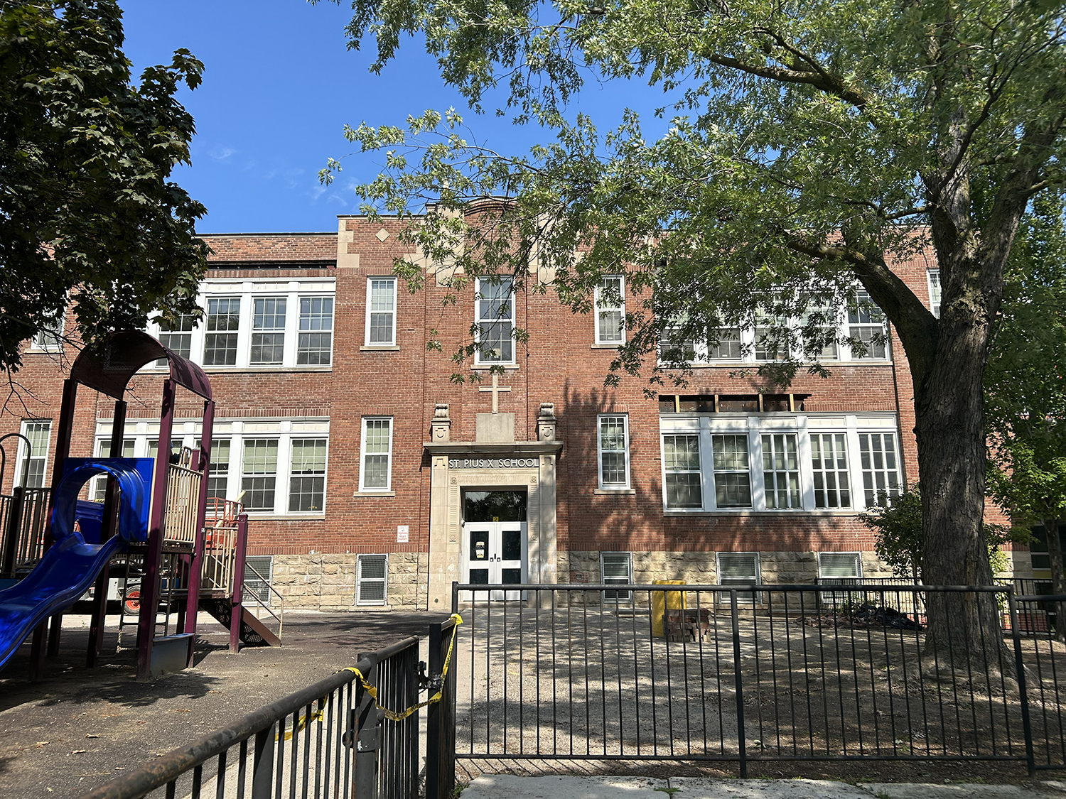 The front of the St. Pius X Catholic School building, showing a light brown building with the school play ground in front and tall trees bordering