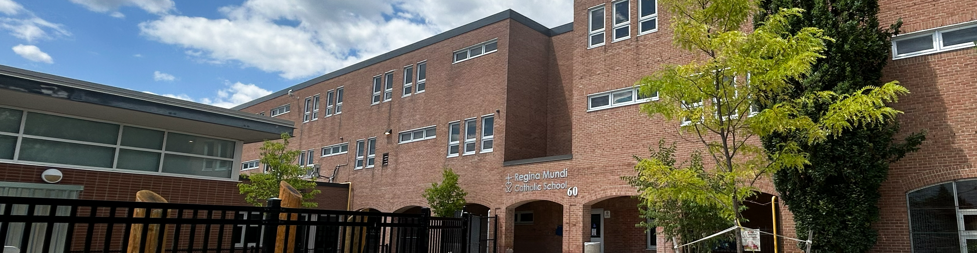 The front of the Regina Mundi Catholic School building, showing a sunlit light brown brick building  bordered with trees, with the school playground in front of it