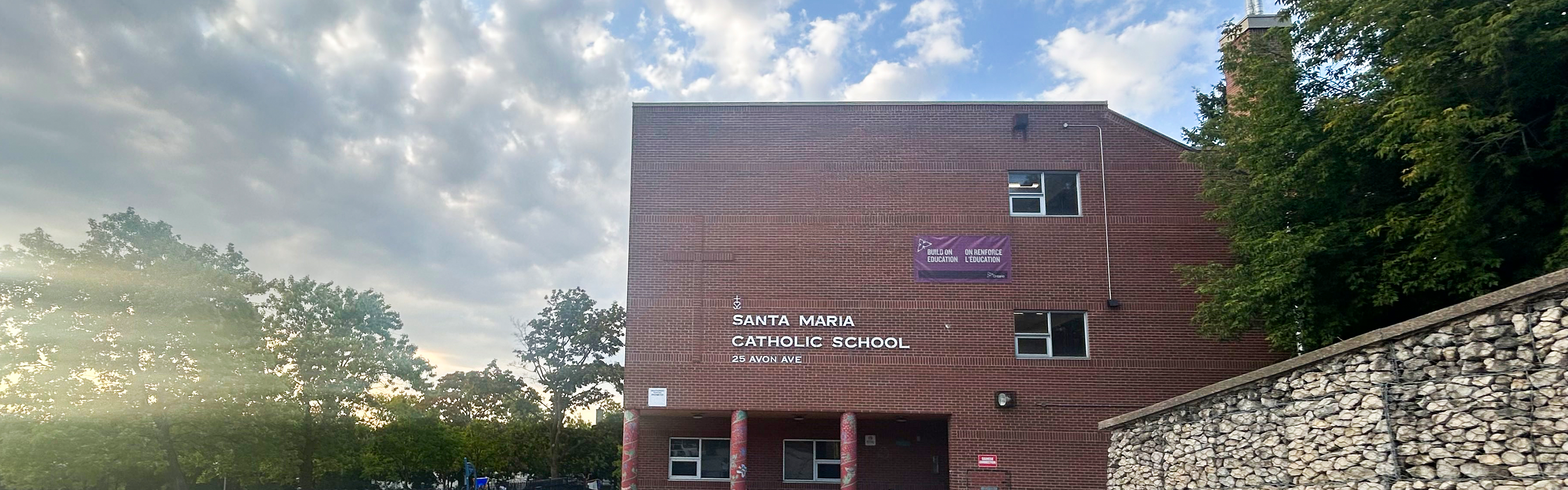 Photo of the Santa Maria Catholic School building, a red brick building with the school name in white on the front, against a vibrant blue and yellow sunset background