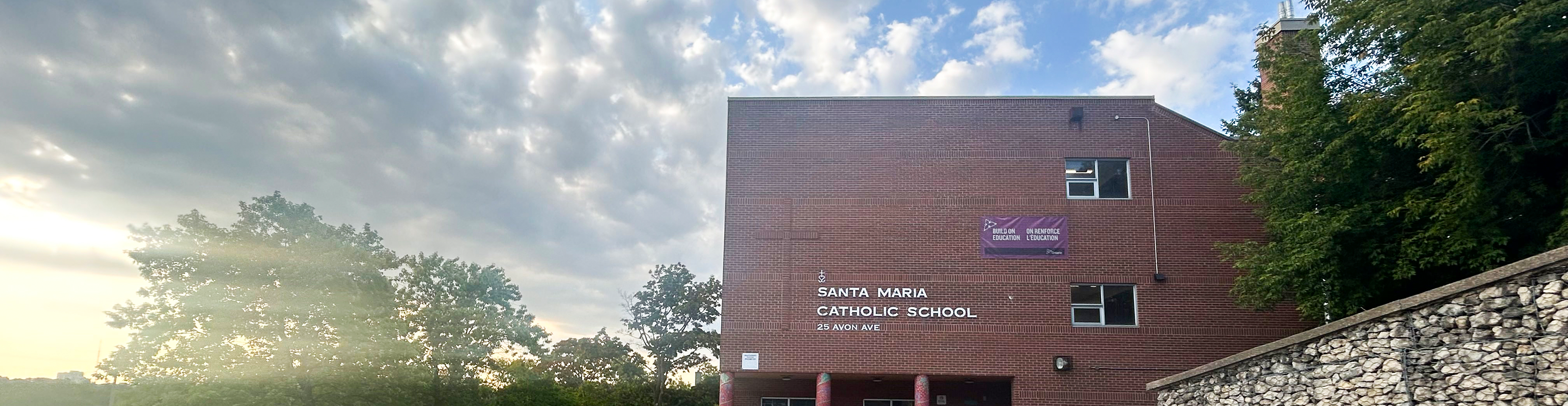 Photo of the Santa Maria Catholic School building, a red brick building with the school name in white on the front, against a vibrant blue and yellow sunset background