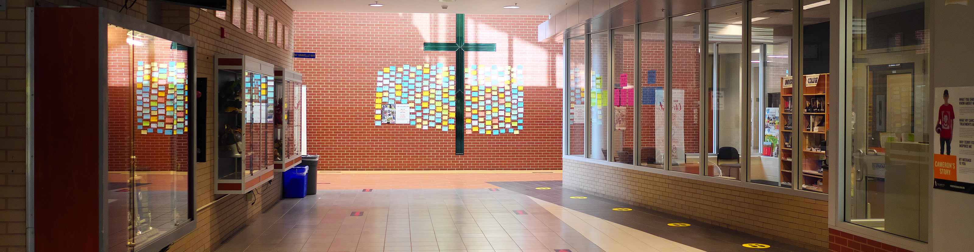 Senator O'Connor school hallways with trophy cases and student work displays on the left side of the hallway, a large green cross on the far wall, and the library on the right side of the hallway.