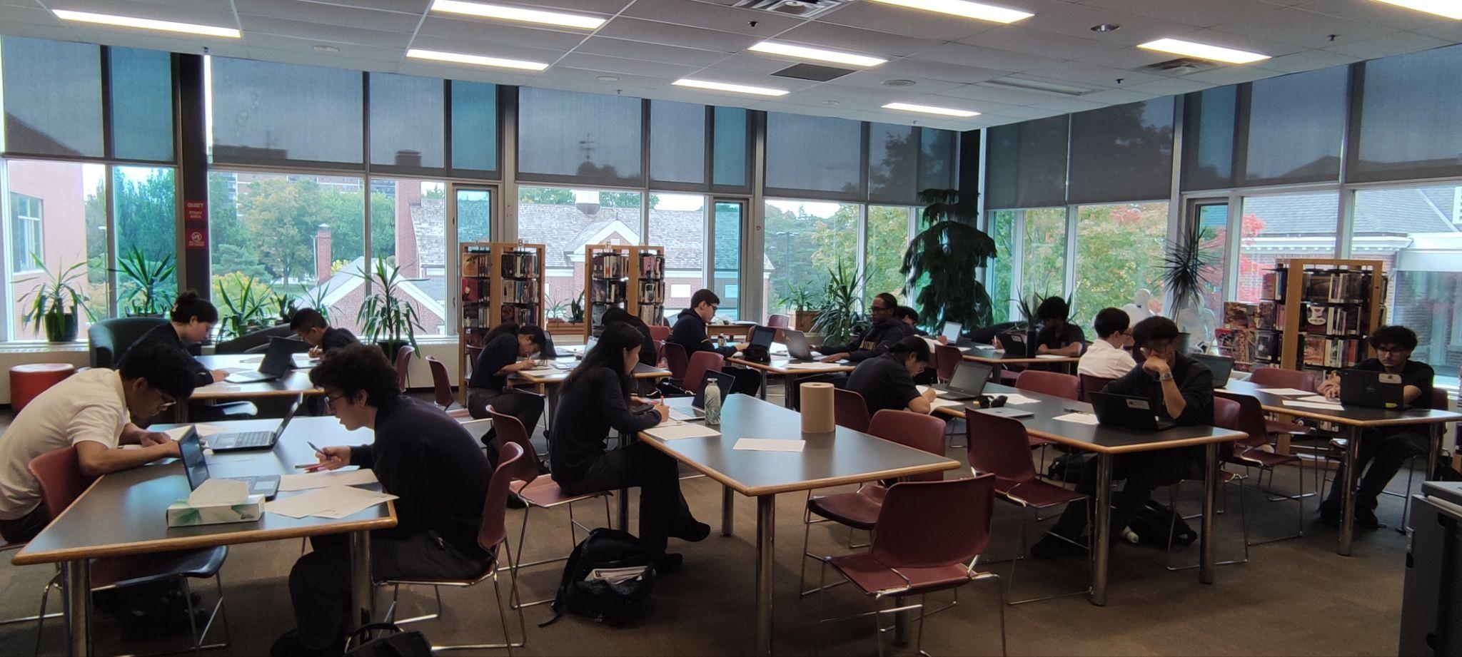Students study and work in groups at tables in a bright library, surrounded by bookshelves and large windows; the atmosphere feels calm and focused.