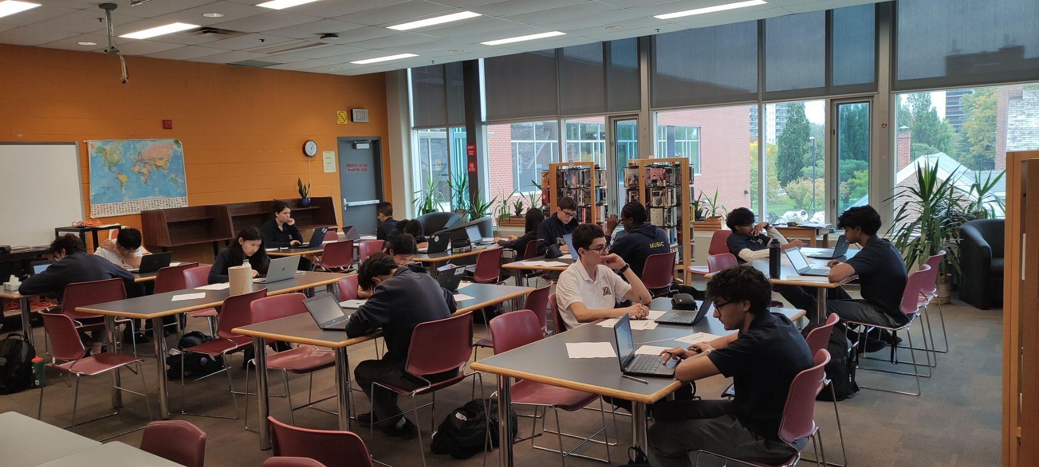 Students study and work on laptops at tables in a bright library, with large windows, bookshelves, and a calm, focused atmosphere.