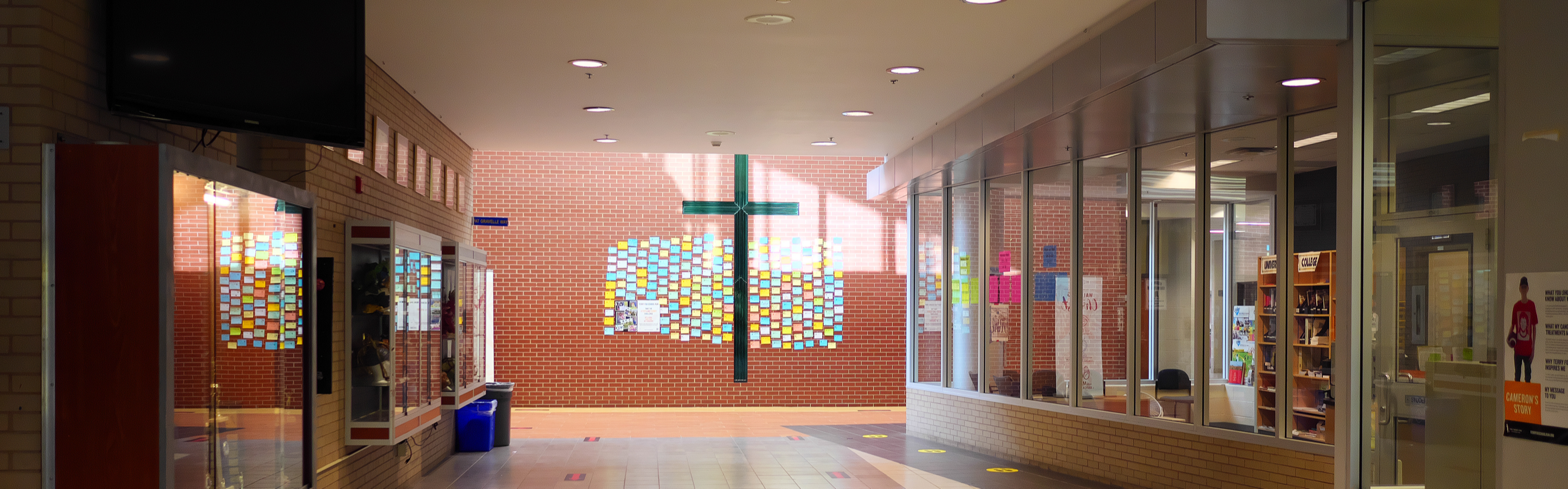 Photo taken from the main entrance of Senator O'Connor College School, the main office to the right and a wall of trophy cases to the left lead up to a sun-lit main lobby featuring a red-brick wall with a large green stained glass window in the shape of a cross, with many colourful papers attached to the wall in a decorative manner.