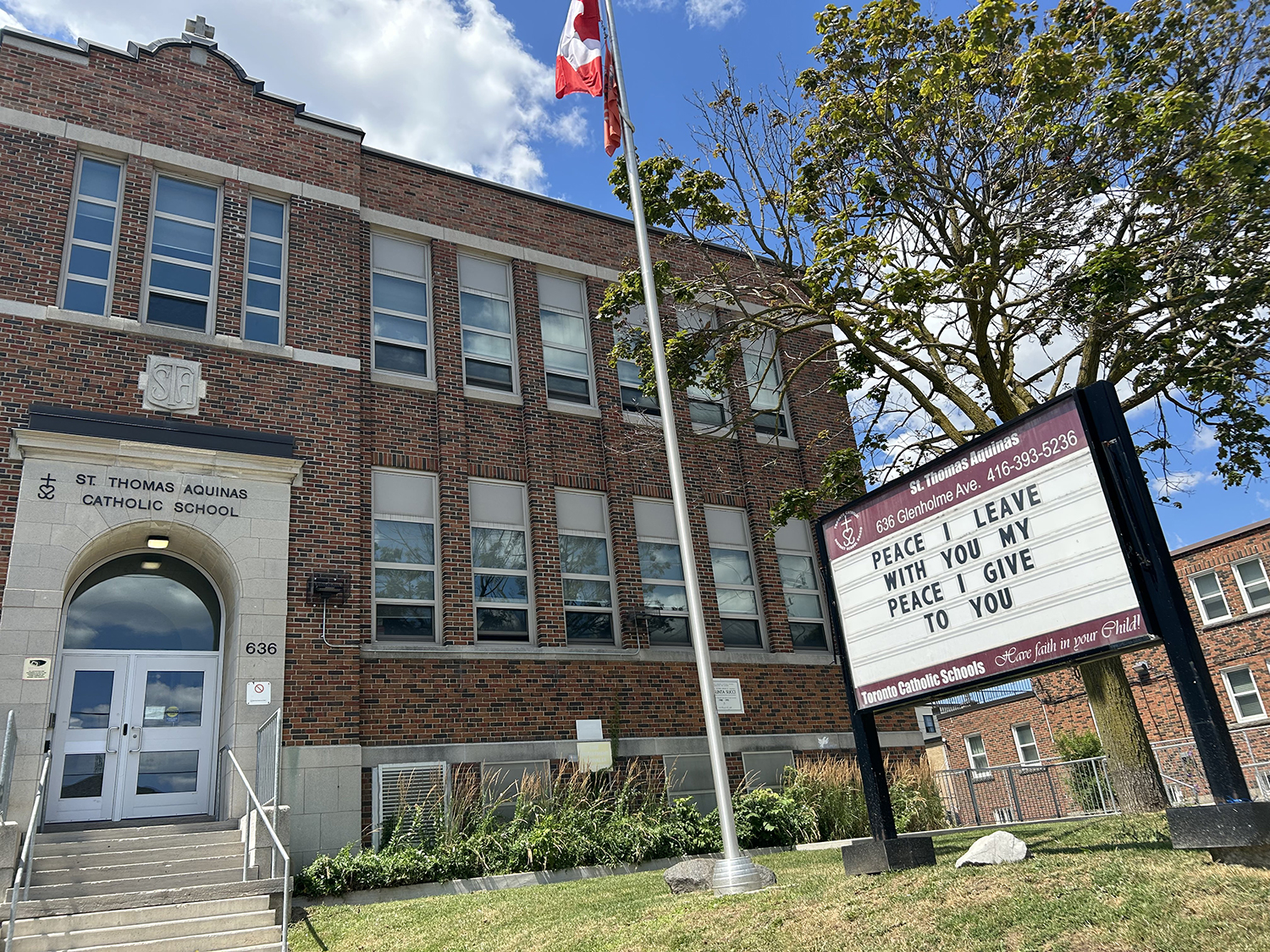 The front of the St. Thomas Aquinas Catholic School building, showing a red brick building with a white stone front, on which the school name is inscribed in black. The building looks out onto the playground and a residential road.