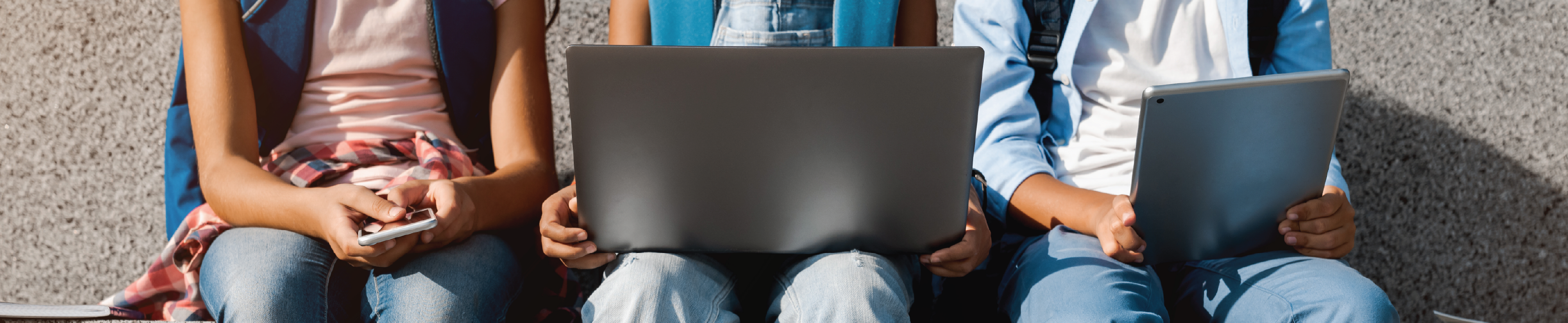 Three students sitting and looking at their phone, laptop and tablet. 