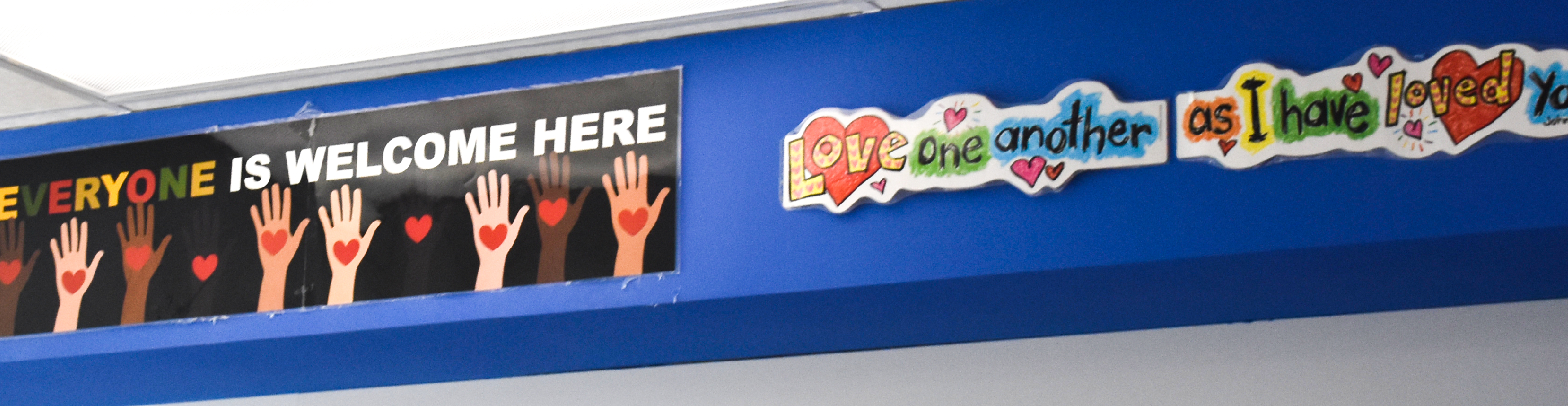 Photo of a school hallway with a banner saying "EVERYONE IS WELCOME HERE" on a background of raised hands of different skin tones, each of them holding a red heart. Next to it is another colourful banner saying "Love one another as I have loved you."