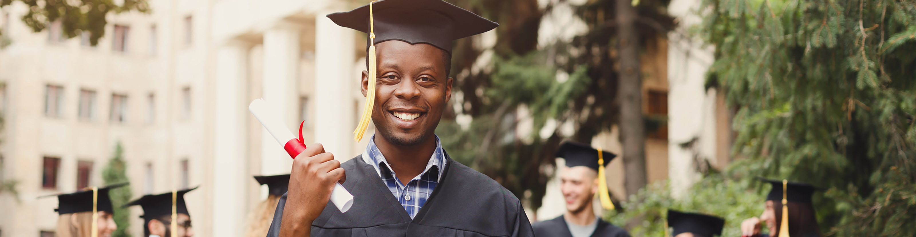 Photo of graduating black student in a graduation robe wearing a mortarboard and holding a diploma, with other students in the background.