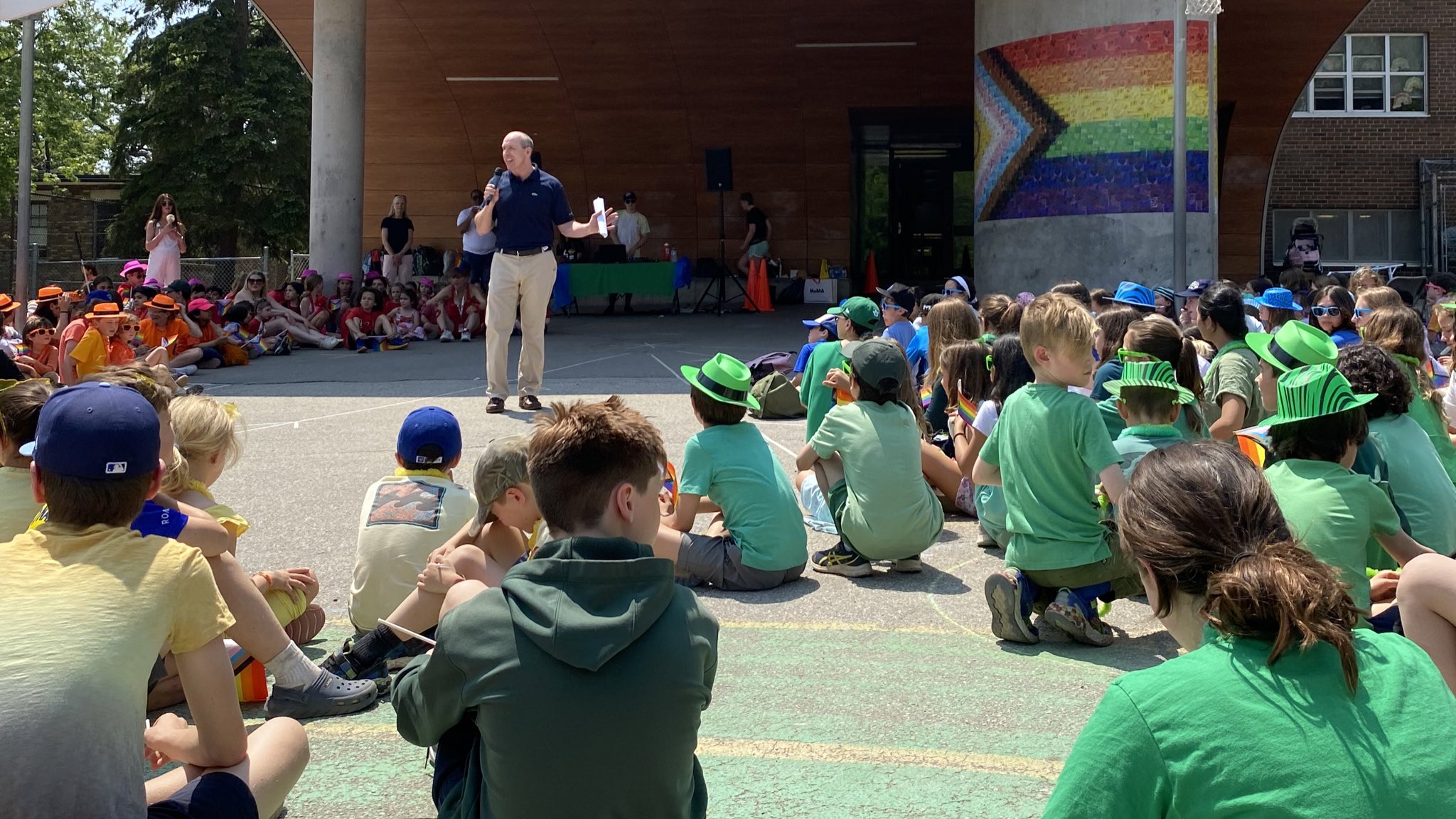 A presenter giving a talk to students in the schoolyard with a Pride flag behind them.