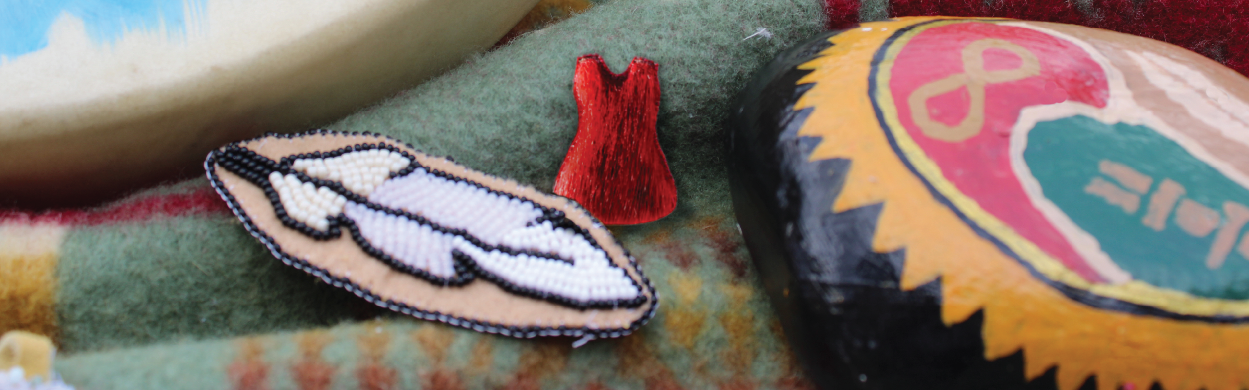 Close-up of Indigenous cultural items arranged on a textured blanket, including a white beaded feather patch, a small red dress ornament, part of a drum, and a painted stone.