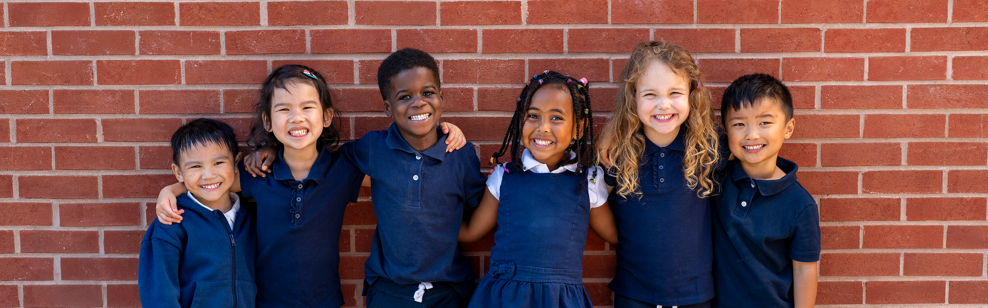 Six elementary school-aged children stand side by side in front of a red brick wall, wearing matching navy blue school uniforms.