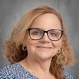 A woman with short blonde curls wears black-framed glasses and a blue patterned top. She smiles.