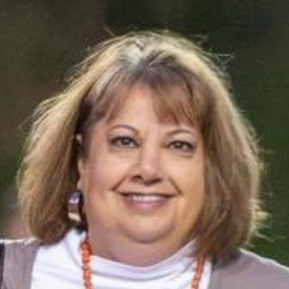 A smiling woman with short, wavy blonde hair and a red beaded necklace. She wears a white turtleneck and has a blurred background.