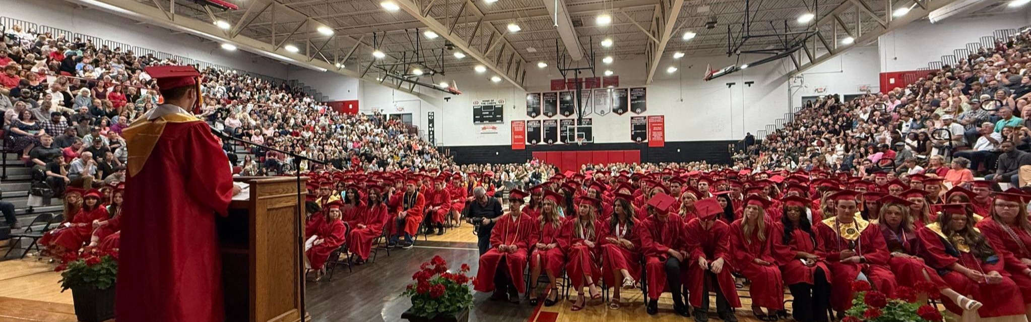 A graduation ceremony in a gymnasium with rows of students in red robes. A person stands at a podium.