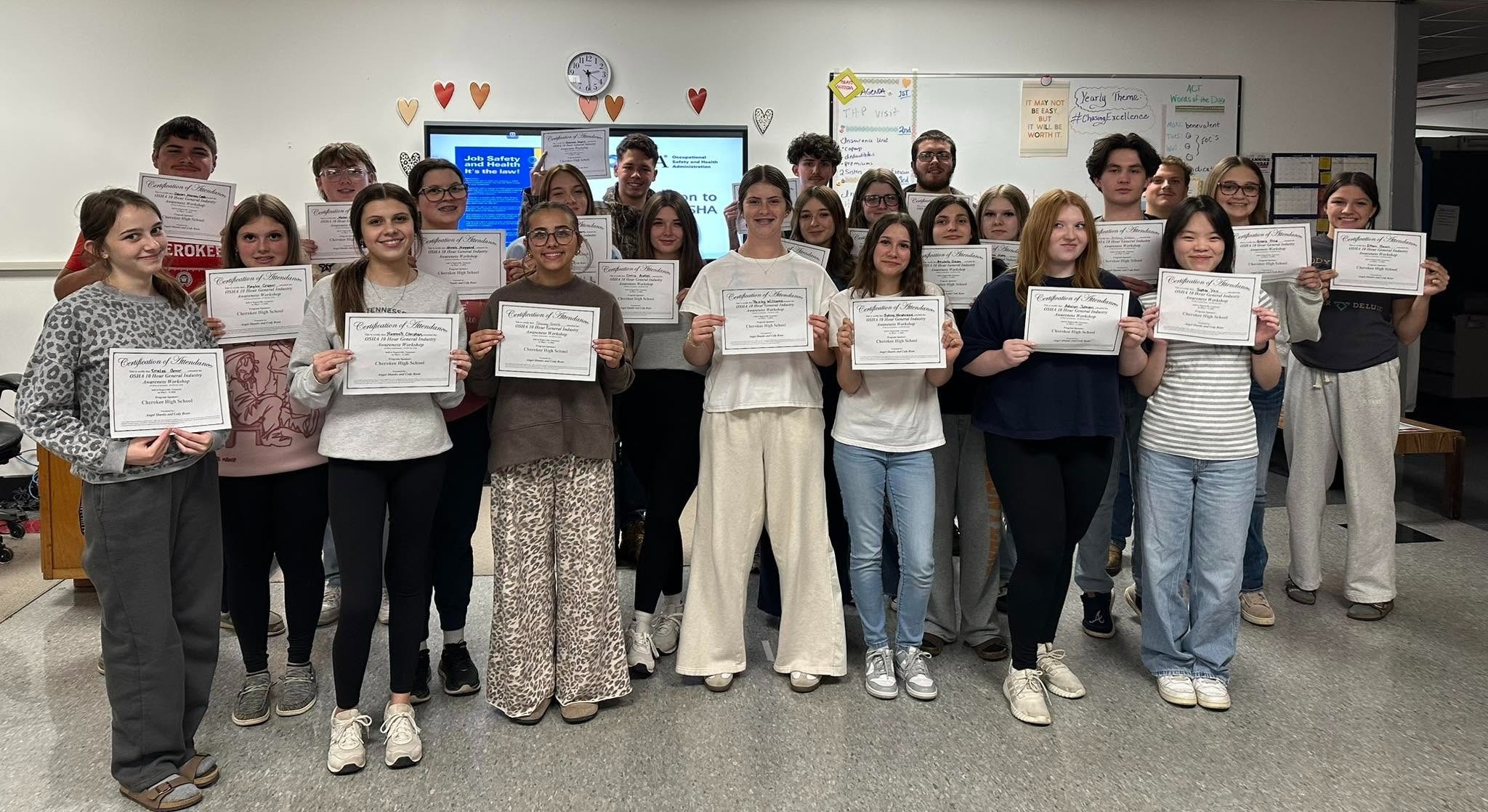 A classroom with a group of students holding certificates in front of a whiteboard and monitors.