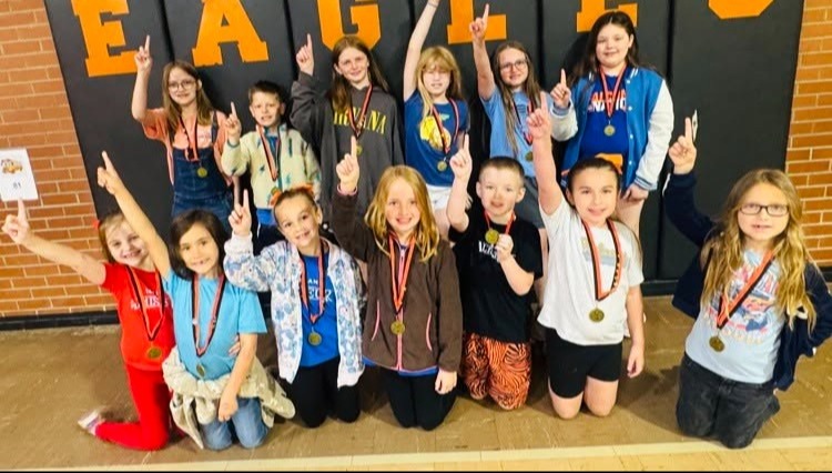 A group of young students, wearing medals, pose for a photo in front of a brick wall with "EAGLE" written on it.